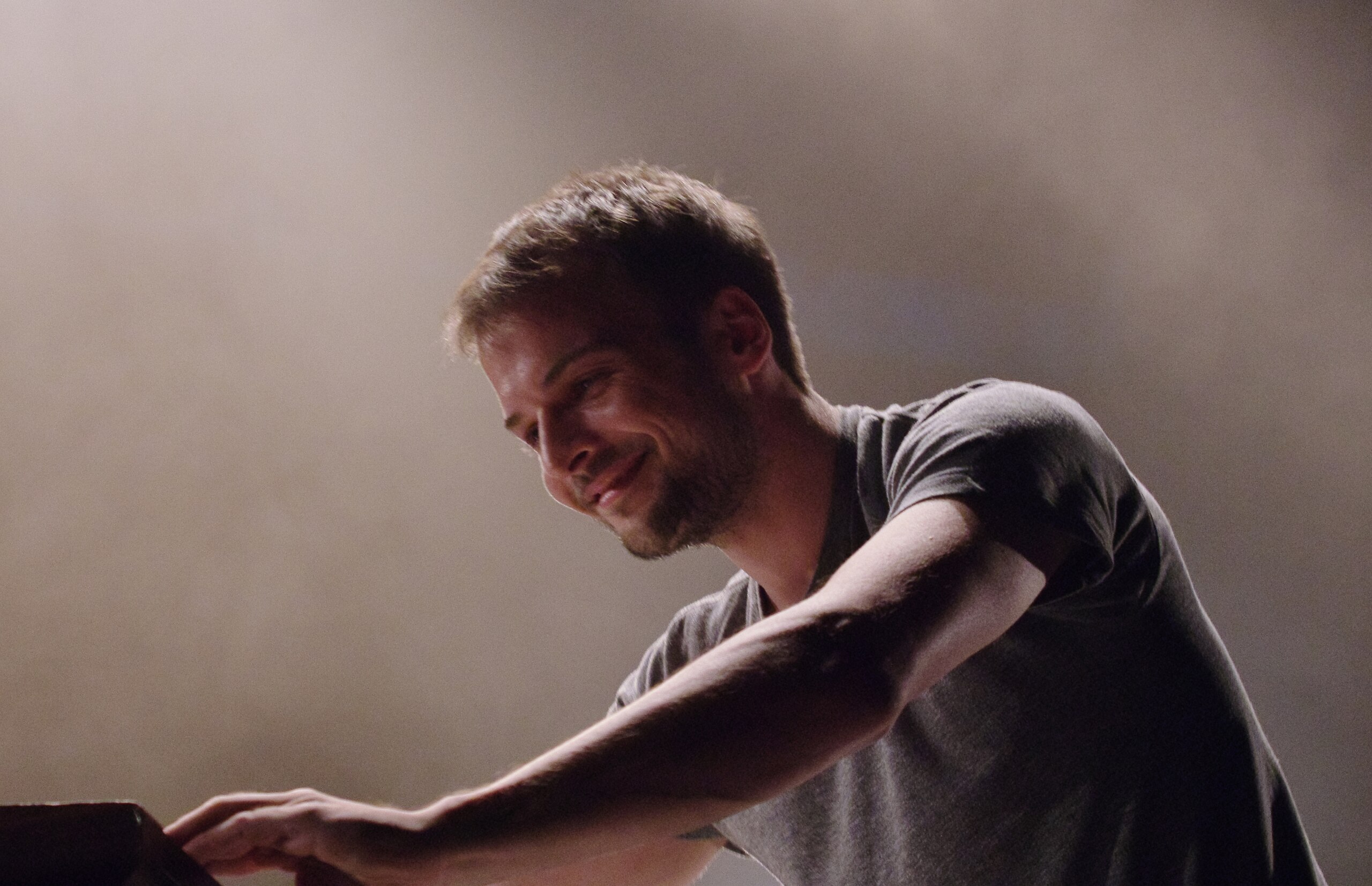 Young man with slight beard smiling while playing piano on stage.
